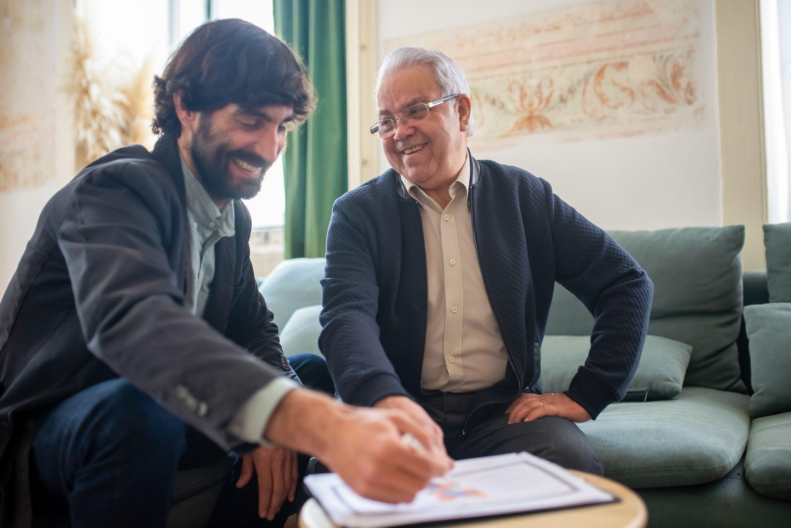 A younger man and an older business owner smiling together while reviewing a document on a coffee table in a relaxed living-room-style office.