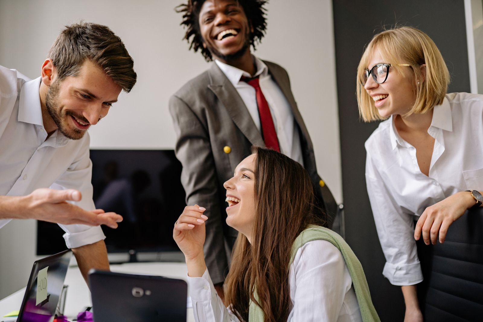 A small office team of four laughing and smiling together while gathered around a laptop.