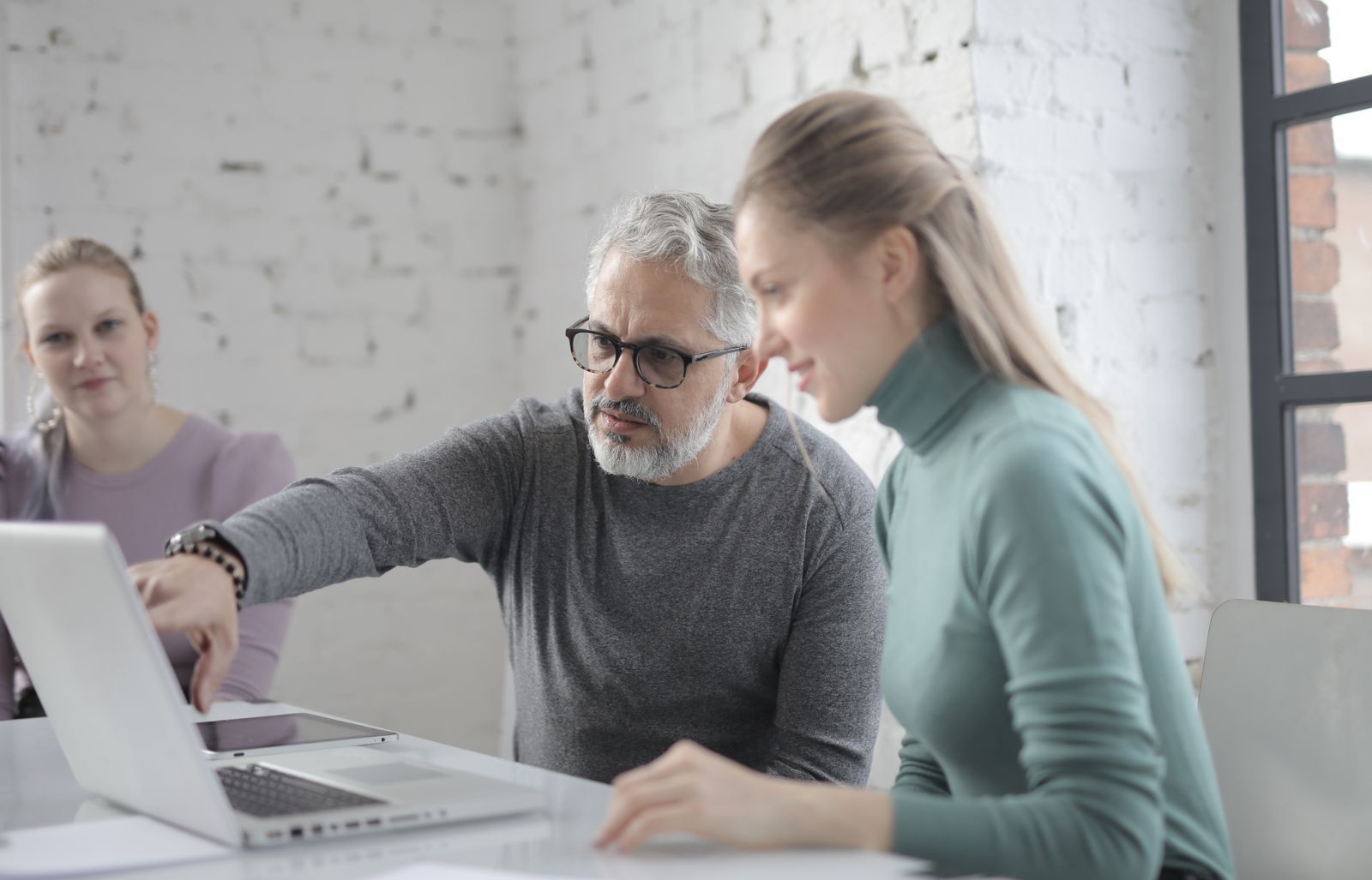 A mature, gray-bearded man pointing at a laptop screen while explaining something to two younger female colleagues in a bright brick-walled office.